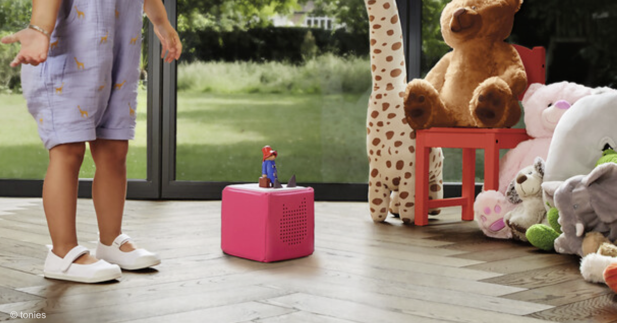 Eine tonie Box steht zwischen einem Kind und Kuscheltieren auf dem Boden