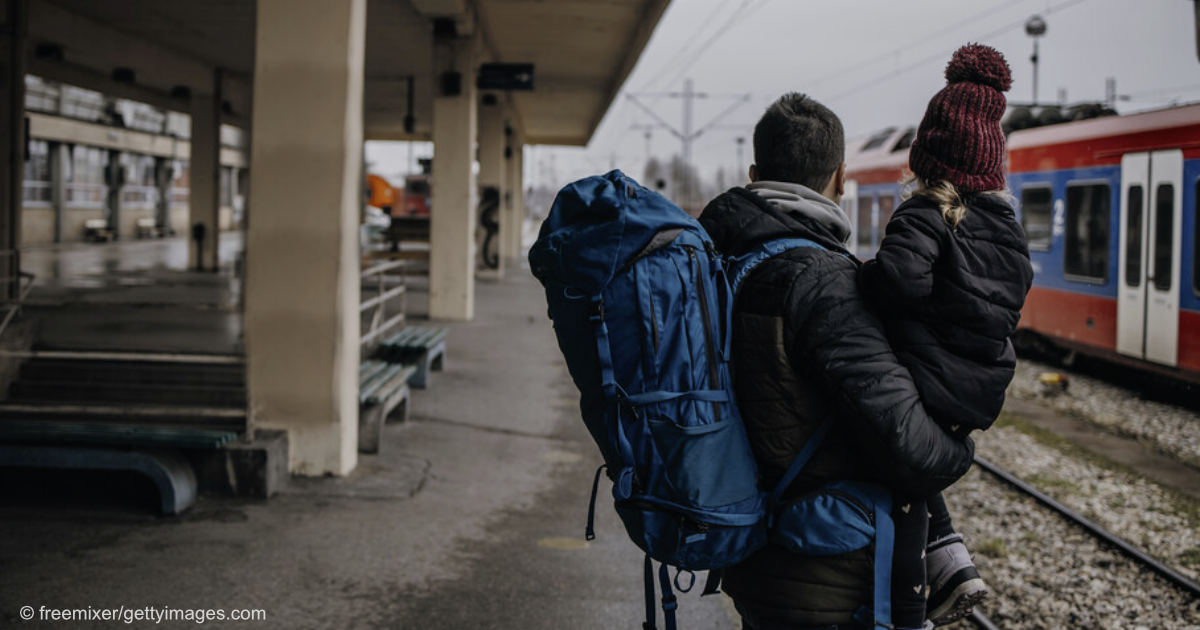 Ein Mann steht an einem Bahnhof und trägt dabei einen Rucksack und ein kleines Kind.