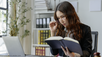 Eine junge Frau sitzt mit einem Buch in der Hand vor einem Laptop